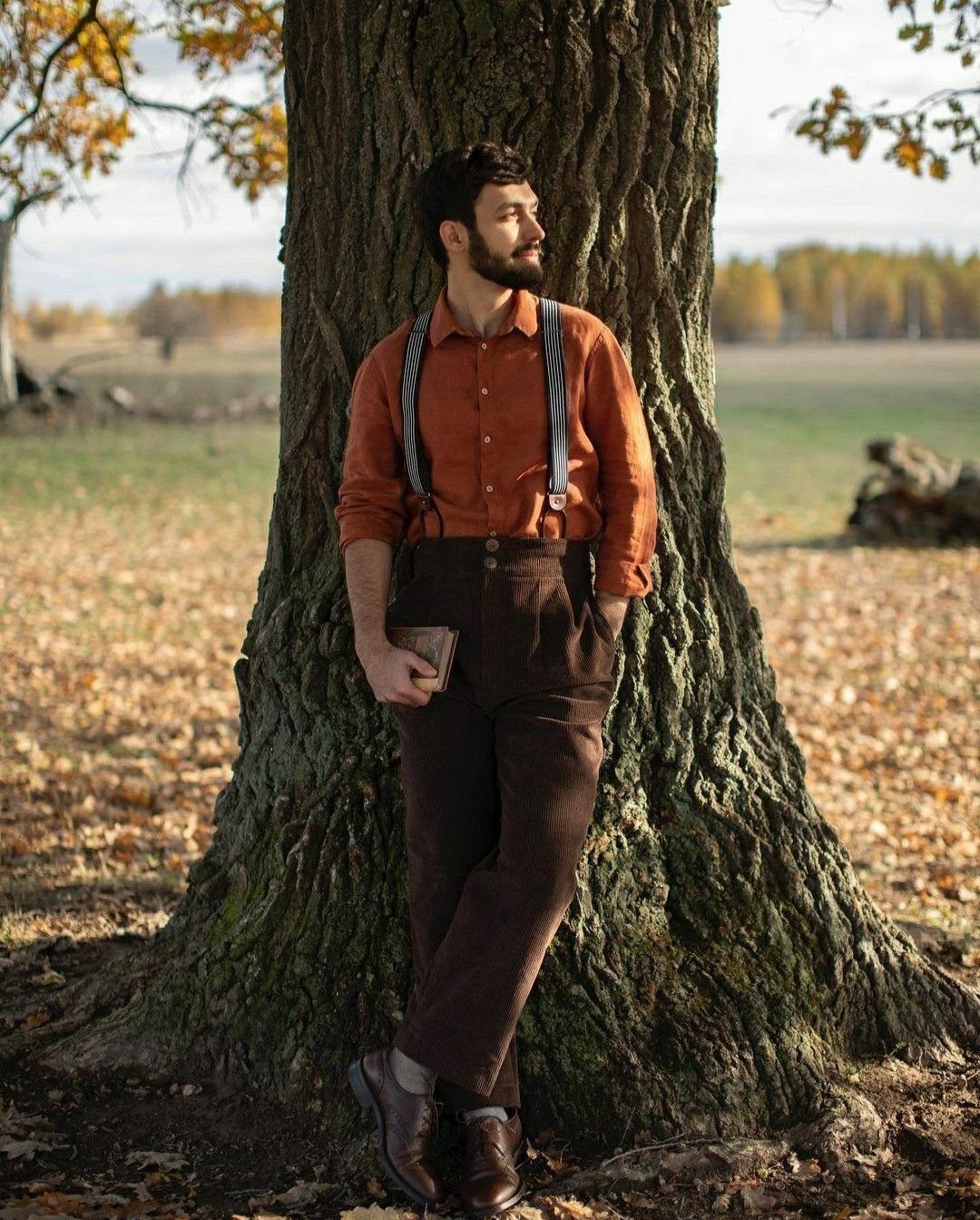 A man in an orange shirt and brown trousers with suspenders leans against a large tree.