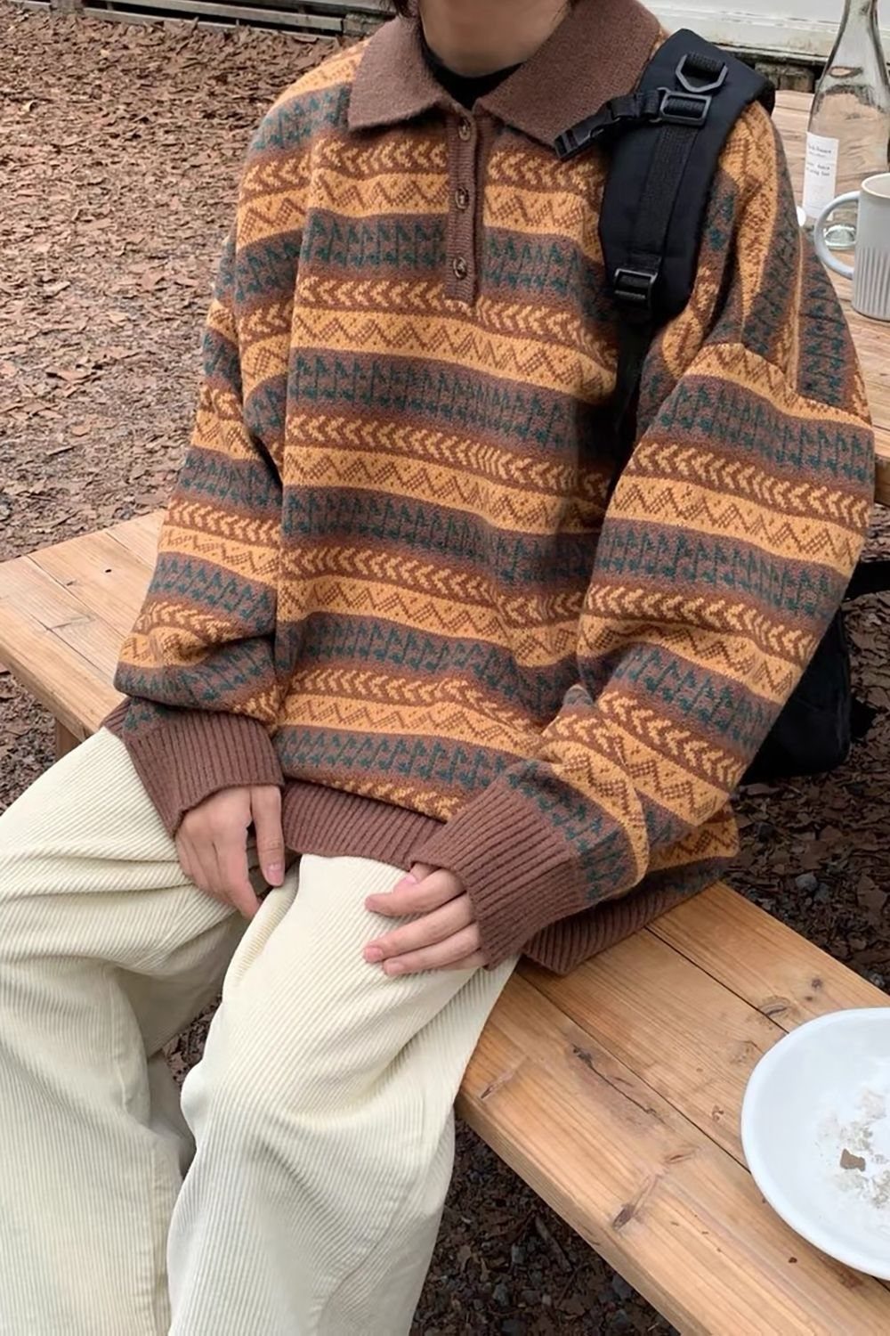 A person wearing a patterned oversized sweater and cream corduroy pants sitting on a wooden bench.