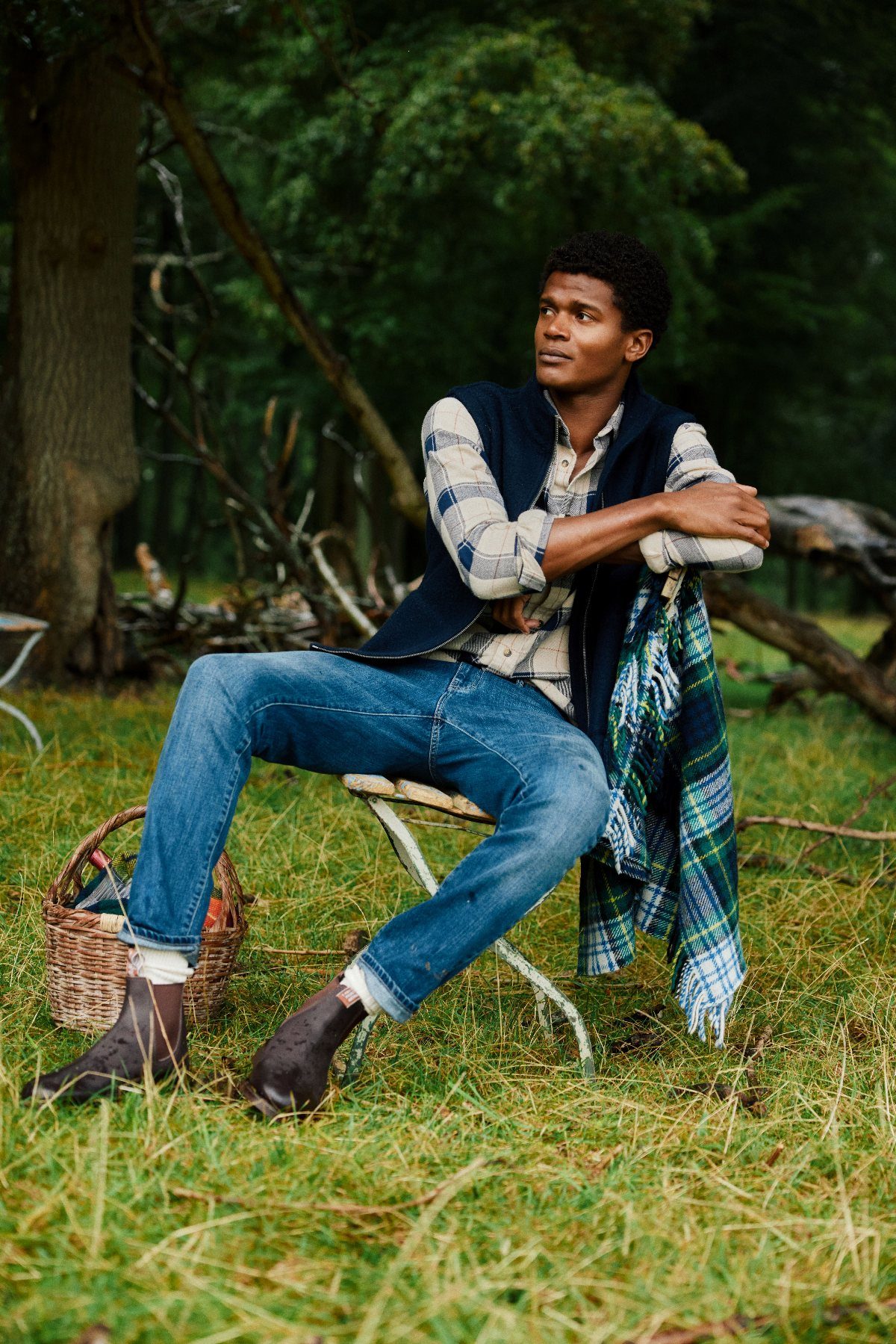 A man wearing a plaid shirt, vest, and jeans sits outdoors in a grassy field.