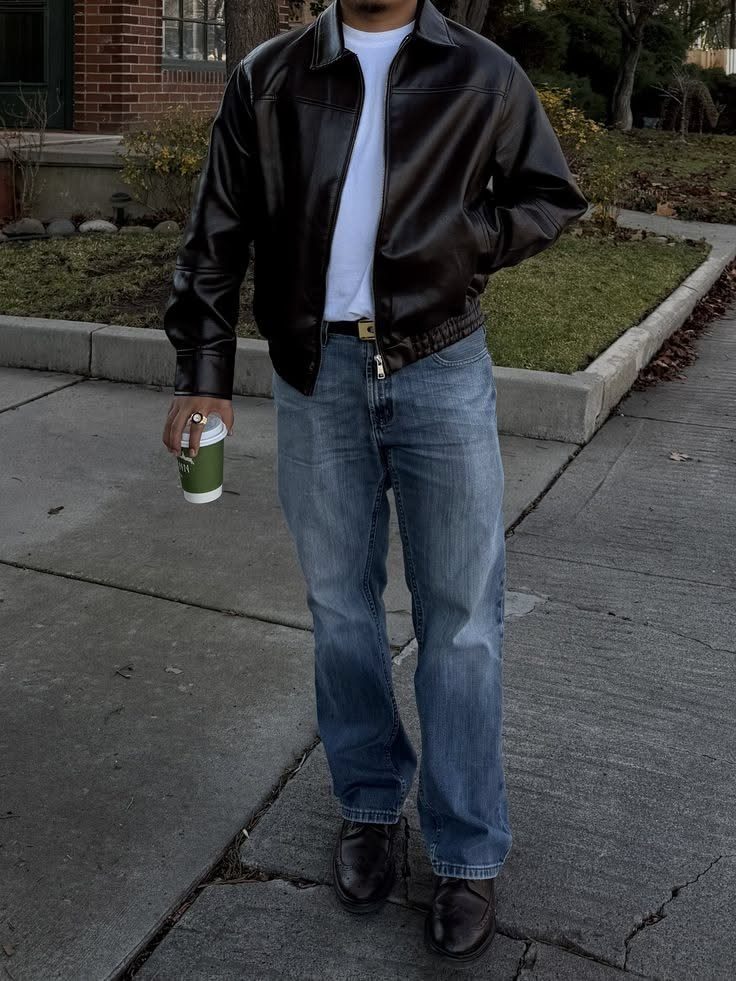 A person wearing a black leather jacket, white t-shirt, and blue jeans standing on a sidewalk.