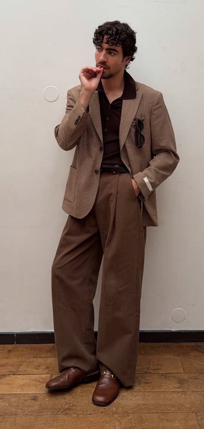 A man posing in a brown blazer, dark polo, wide trousers, and leather loafers.