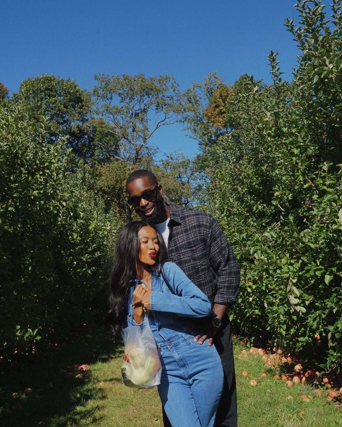 A couple posing together in an apple orchard holding a bag of freshly picked apples.