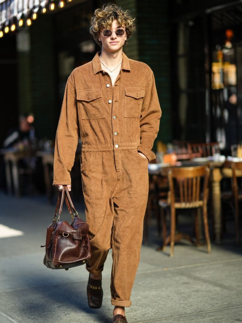 A young man walking in a city wearing a brown corduroy jumpsuit and sunglasses.