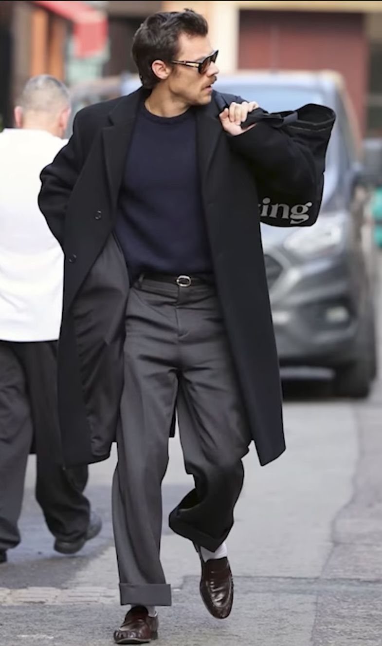 A man walking in a city wearing a black overcoat, navy sweater, and grey trousers.