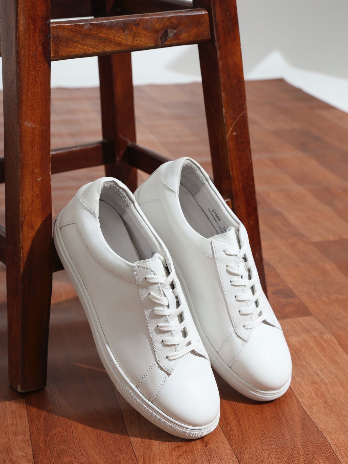 White leather casual sneakers resting on a wooden floor beside a wooden stool.