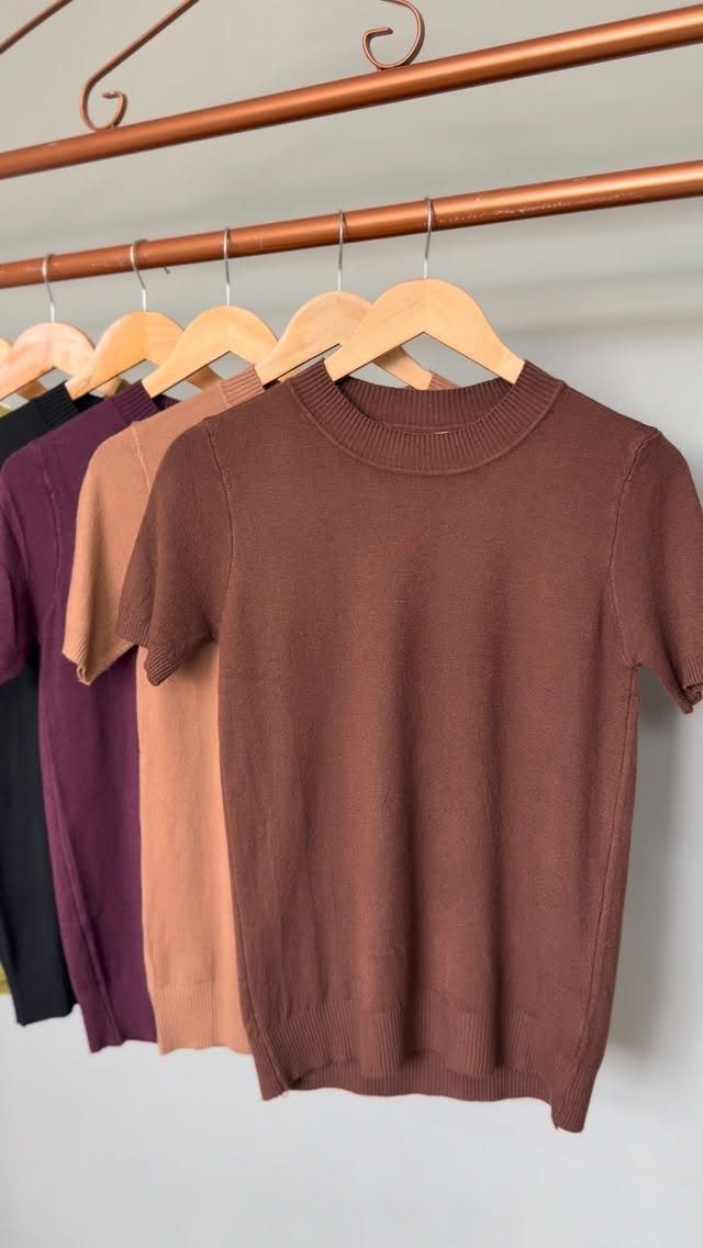 Short-sleeved knit tops in neutral tones hanging on wooden hangers on a copper rack.