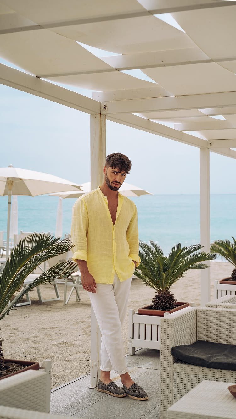 Man in yellow linen shirt and white pants wearing grey espadrilles on a beach.