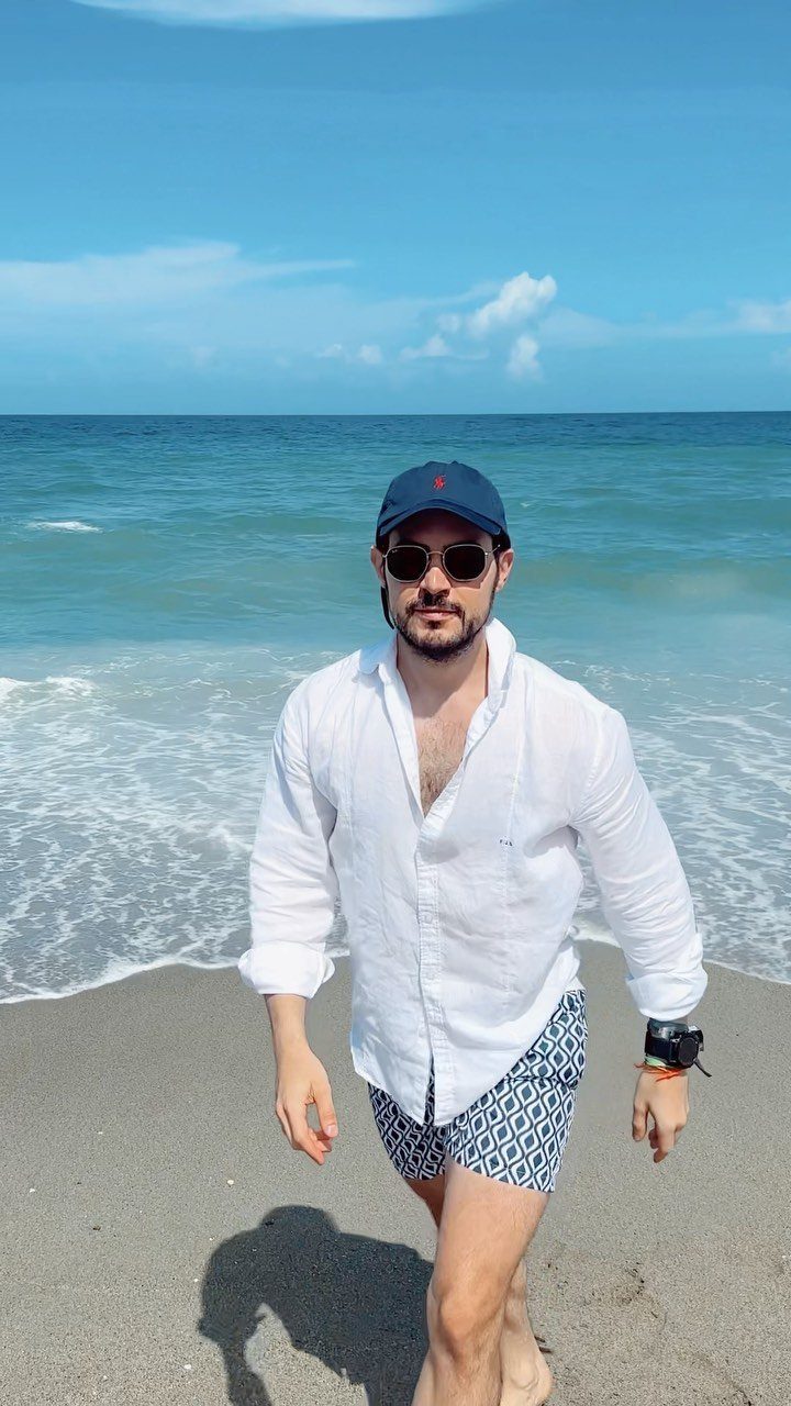 Man in white linen shirt and patterned swim shorts walking on a sunny beach.