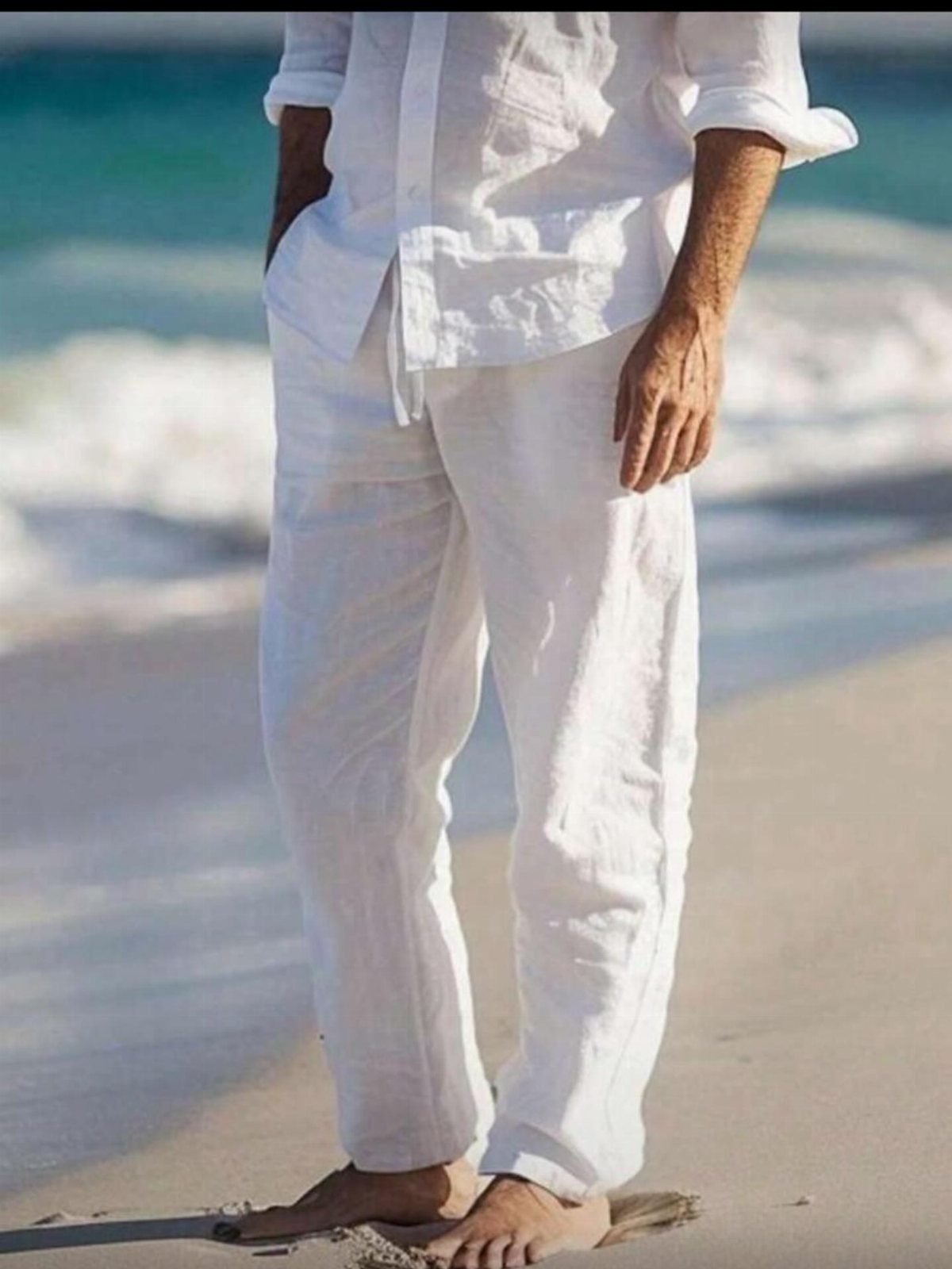 Man wearing white linen pants and a matching shirt standing on a sandy beach.