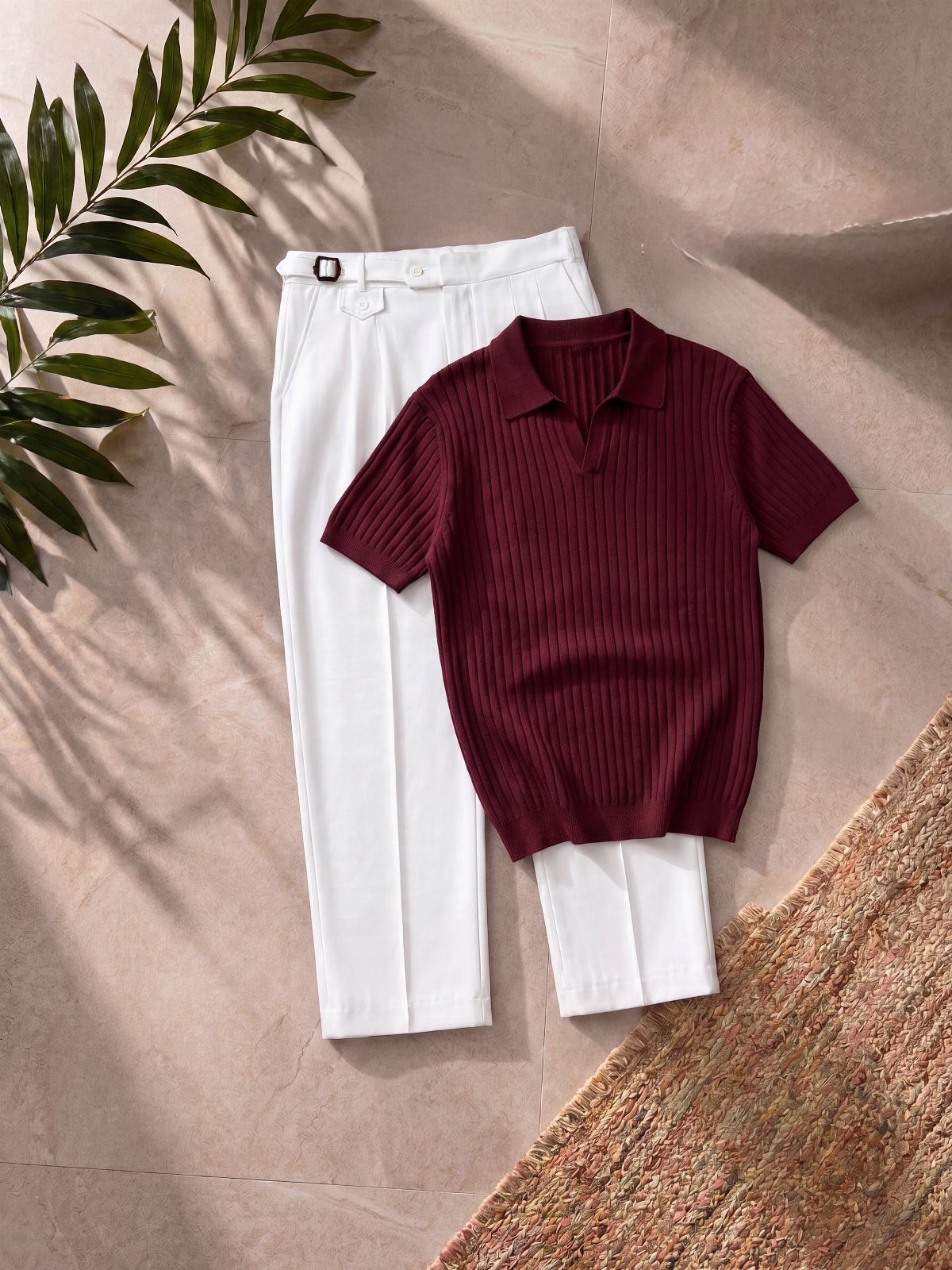 A maroon ribbed polo shirt and white trousers laid on a tan surface with palm leaves.