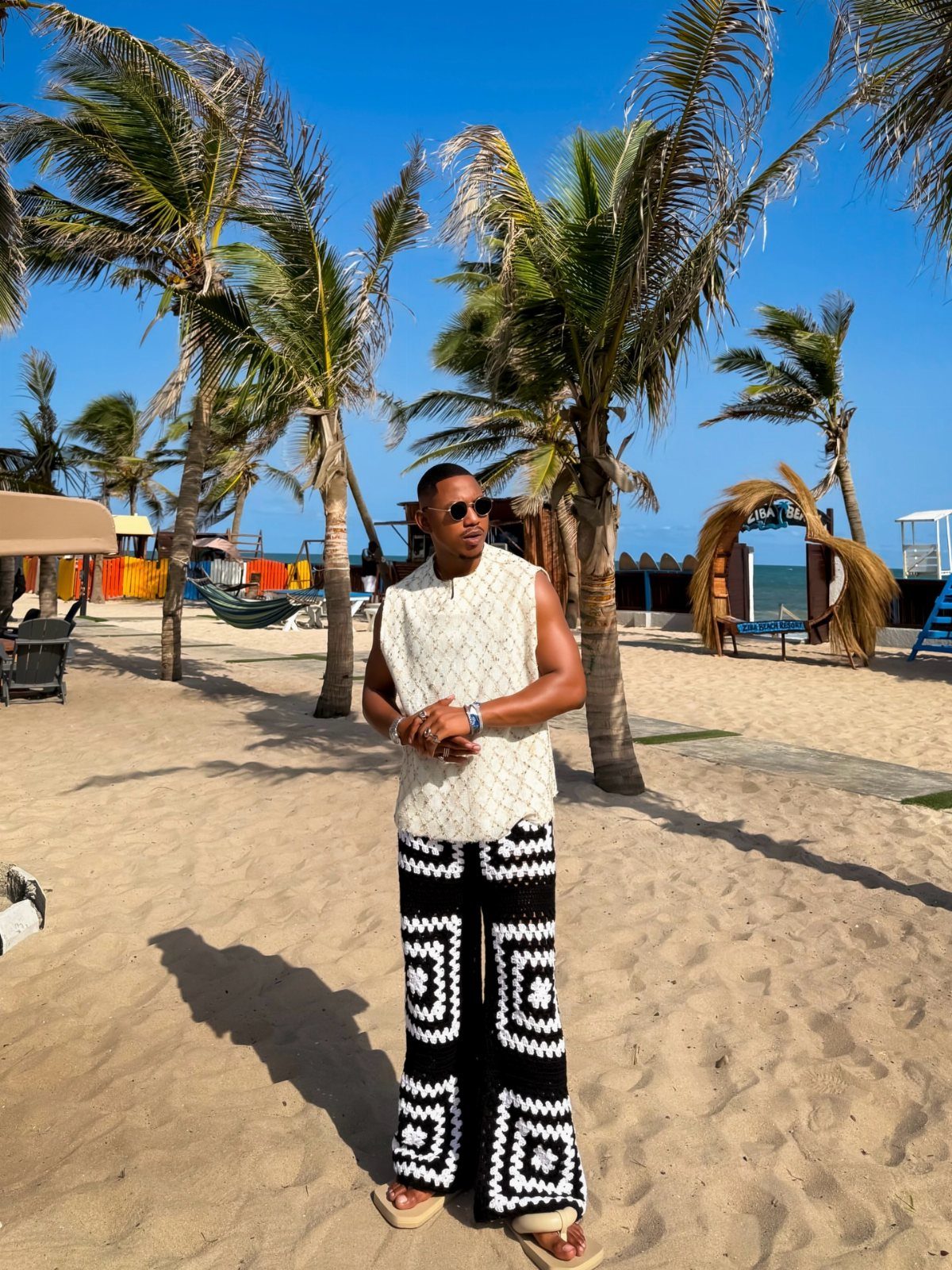 A man in a white textured top and patterned wide-leg trousers standing on a sandy beach.