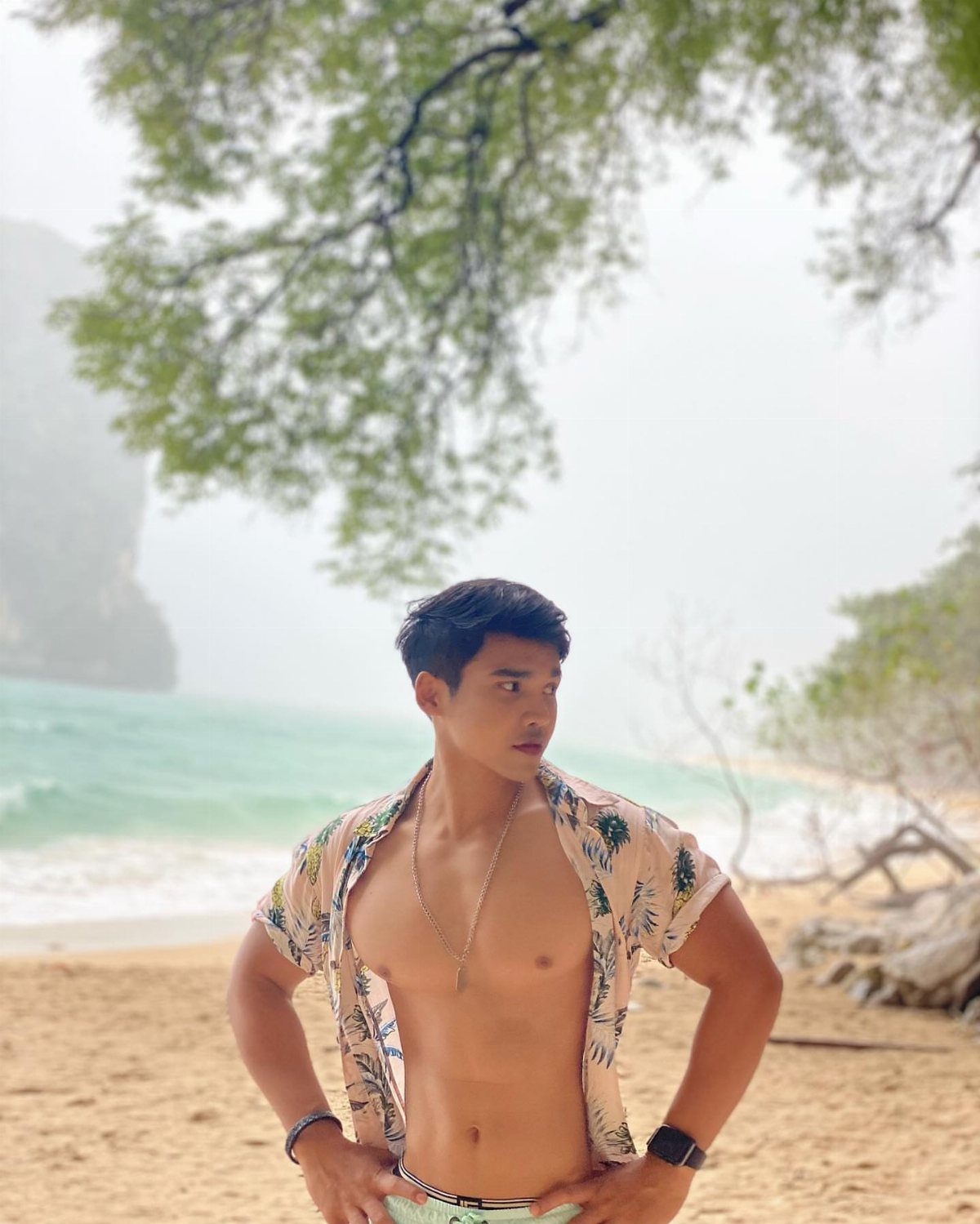 A young man posing on a tropical beach wearing an open floral print shirt.