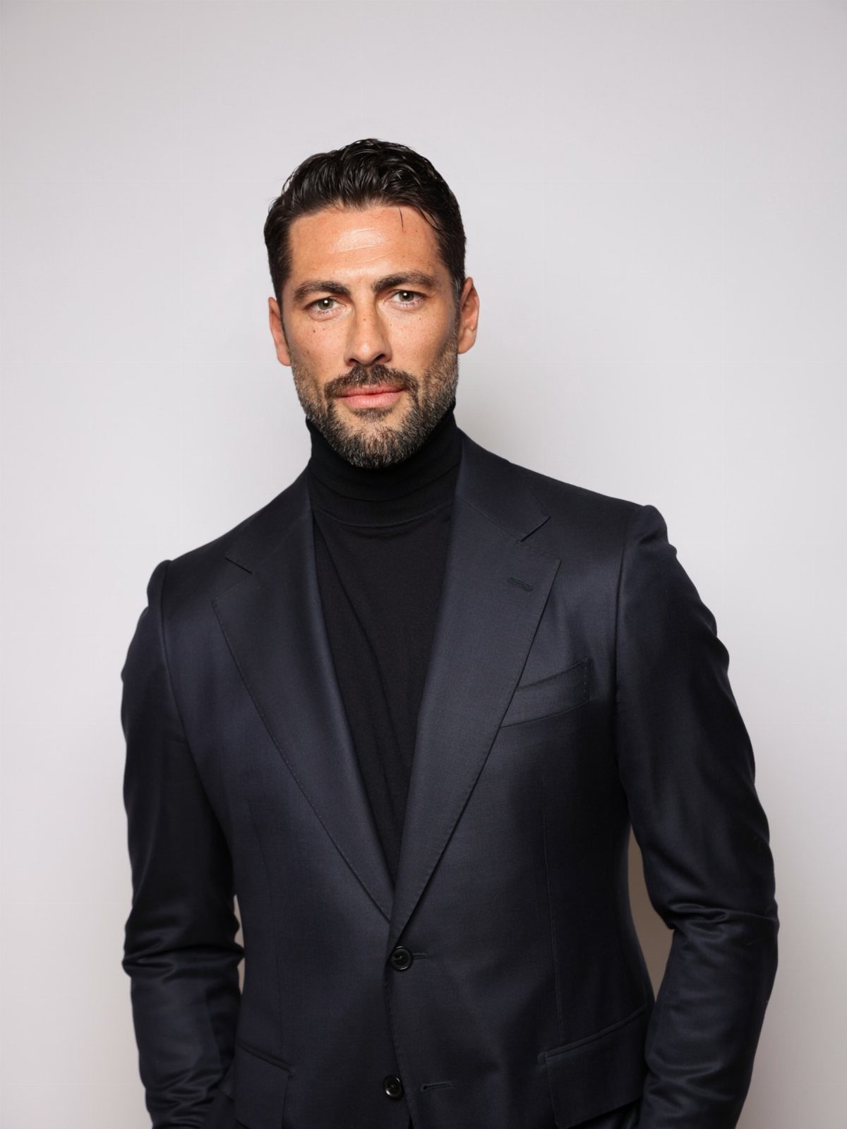 A man wearing a navy blue blazer and a black turtleneck sweater against a grey background.