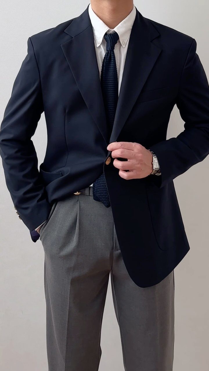 A man dressed in a navy blue blazer, white shirt, dark tie, and gray trousers.