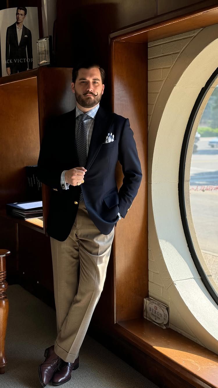 A man wearing a navy blue blazer, light blue shirt, tie, and khaki pants leaning against a window.