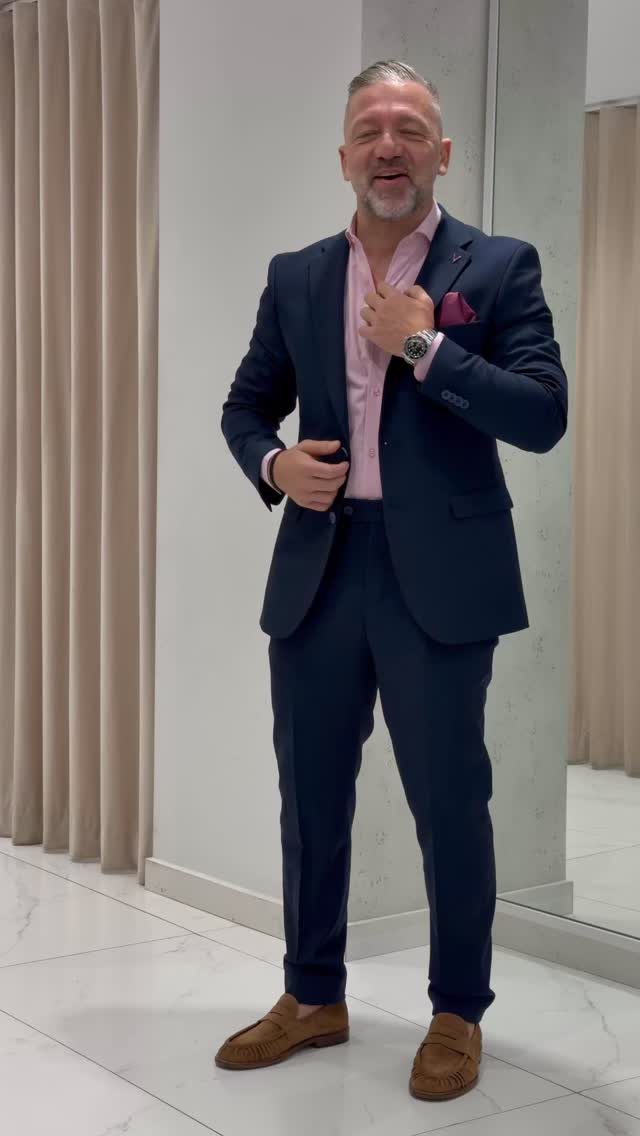 A man smiling while wearing a navy blue blazer, pink shirt, and brown loafers.