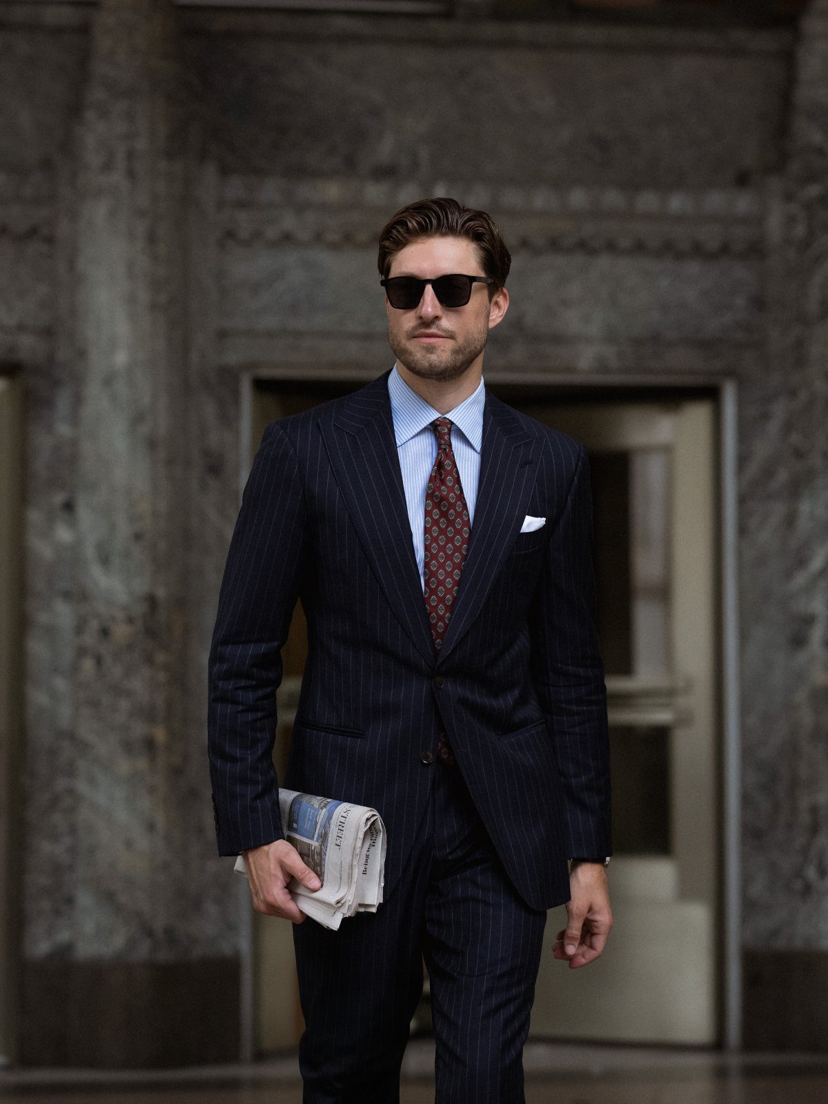 Man in a navy pinstripe suit and light blue shirt walking with a newspaper.