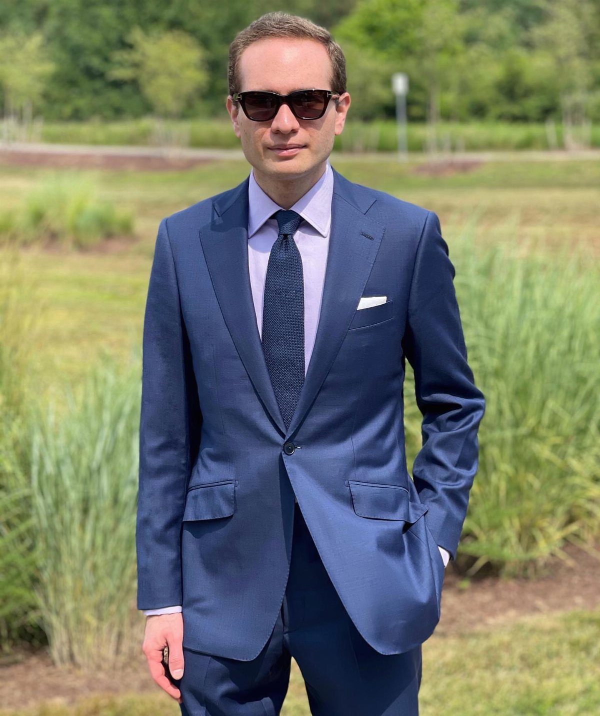 A man in a navy blue suit and lilac shirt standing in a grassy field.