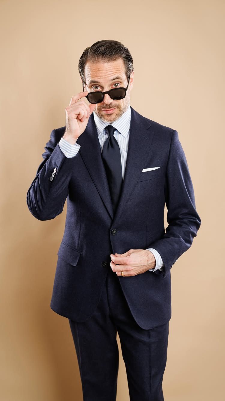 Man in a navy blue suit and striped shirt holding sunglasses against a tan background.