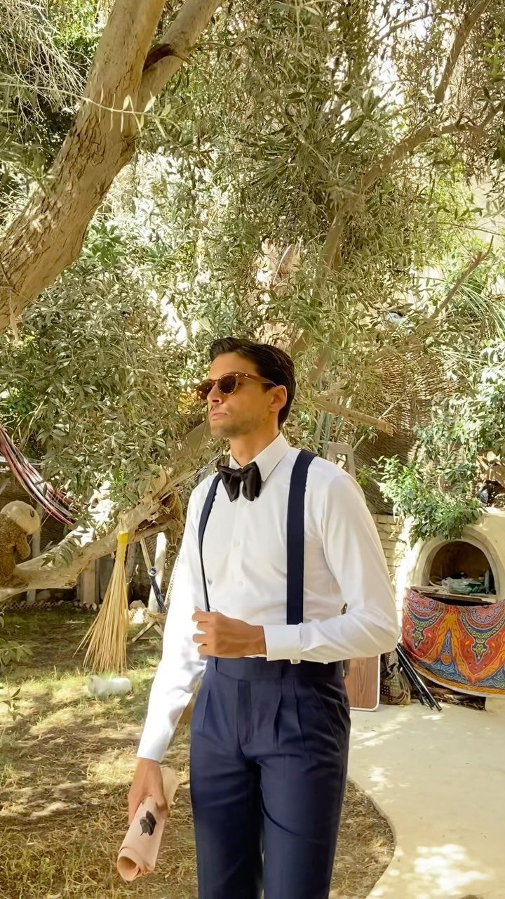A man in a white shirt, black bow tie, and navy suspenders standing under trees.