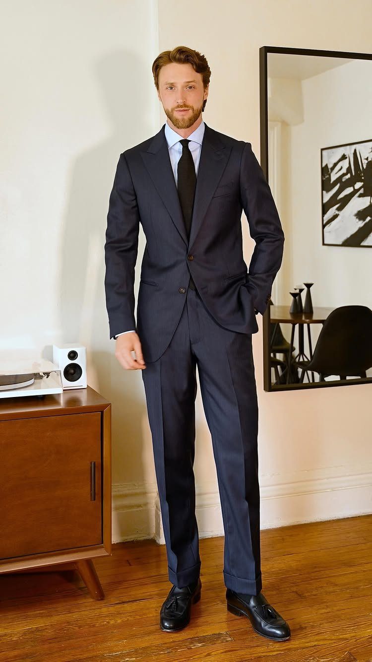 A man in a navy blue suit, light blue shirt, and black tie standing in a room.