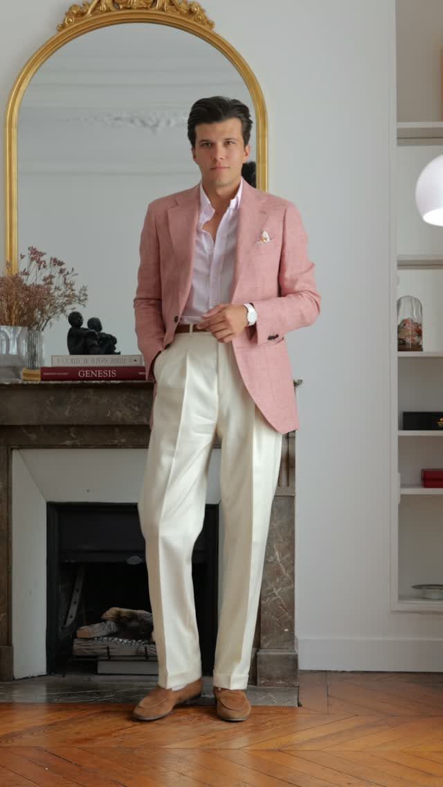 Man wearing a pink blazer, white shirt, and cream trousers standing by a fireplace.