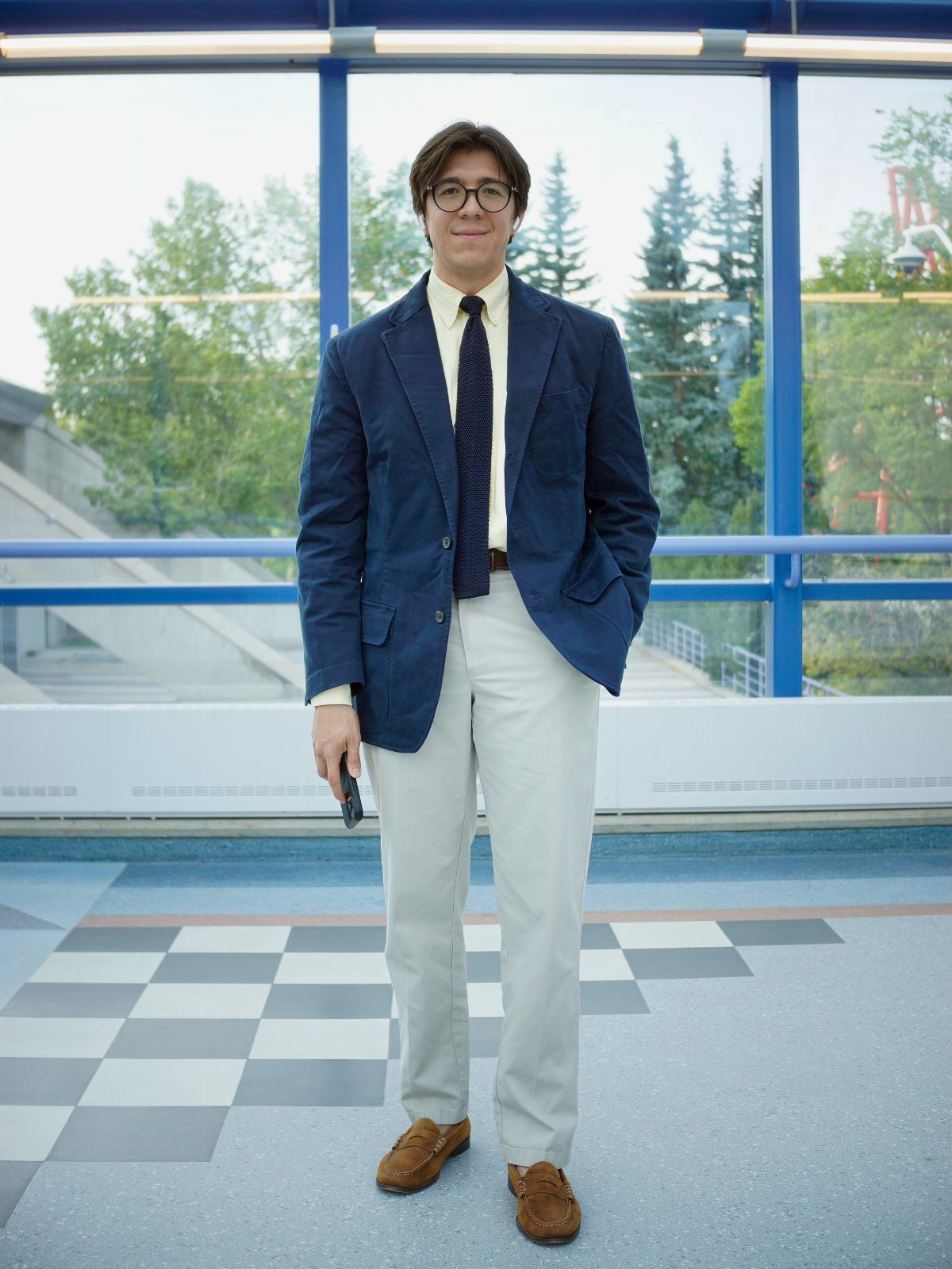 A man dressed in a navy blazer, yellow shirt, tie, and grey chinos standing indoors.