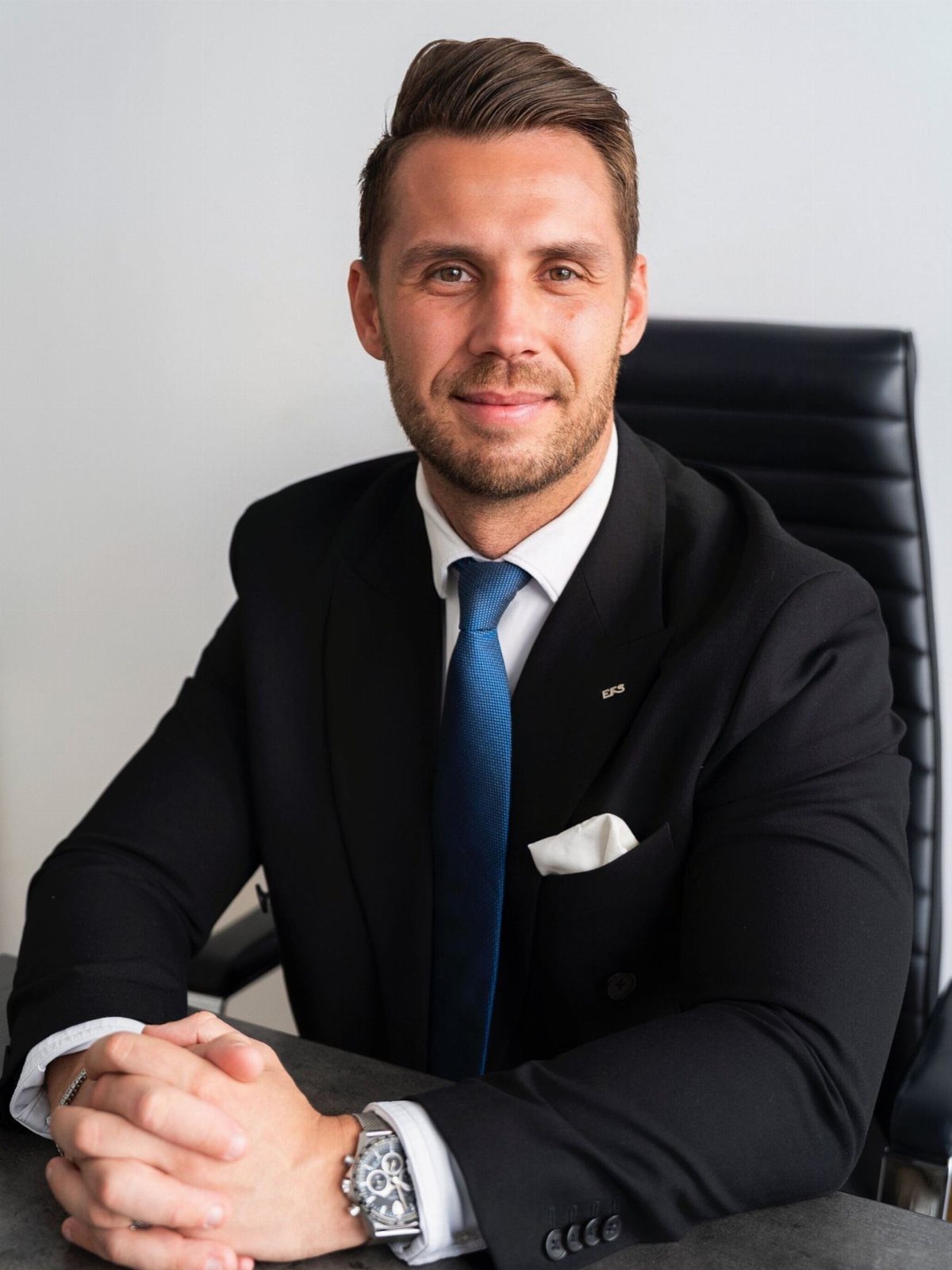 A smiling man in a black business suit and blue tie sitting at a desk.