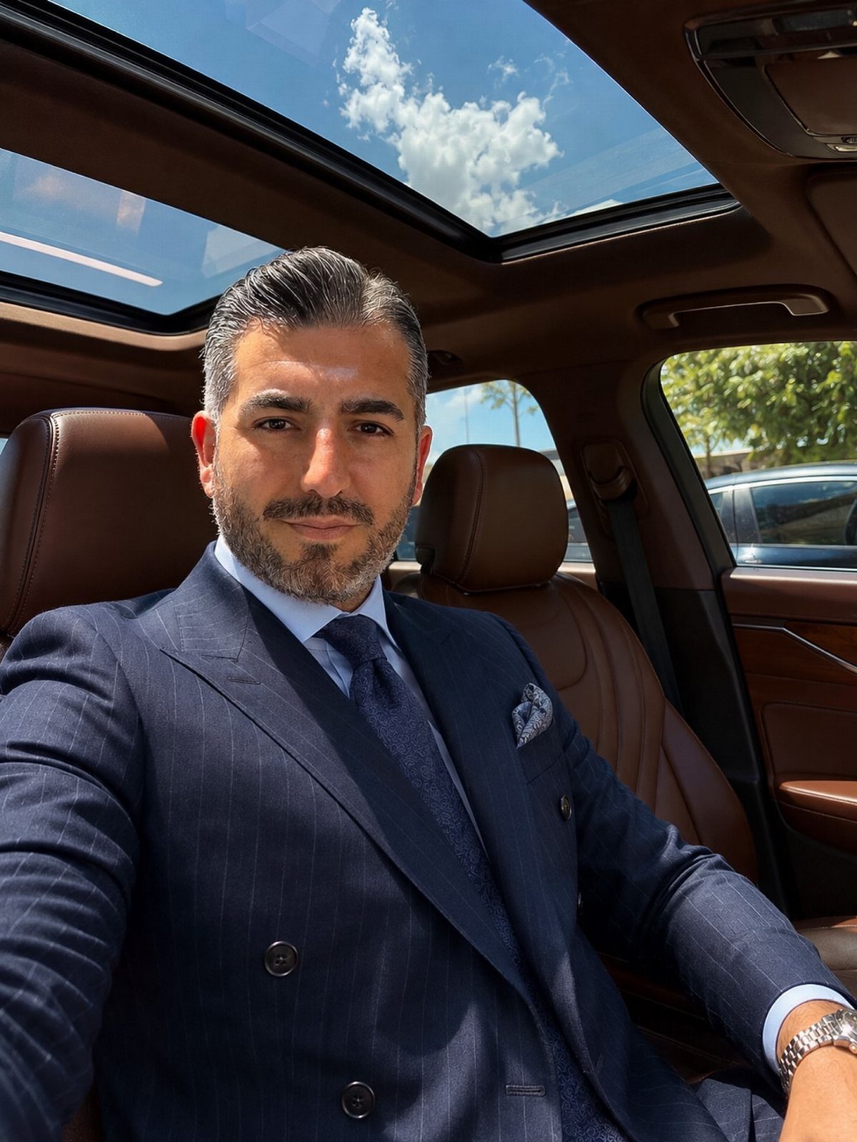 A man with groomed facial hair and styled hair wearing a navy pinstripe suit.