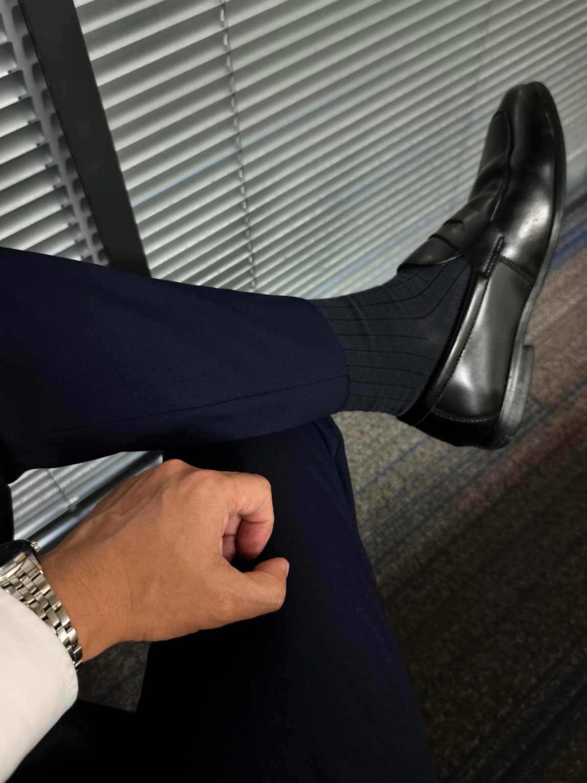 A person wearing navy trousers, black socks, and polished black leather loafers sitting by blinds.