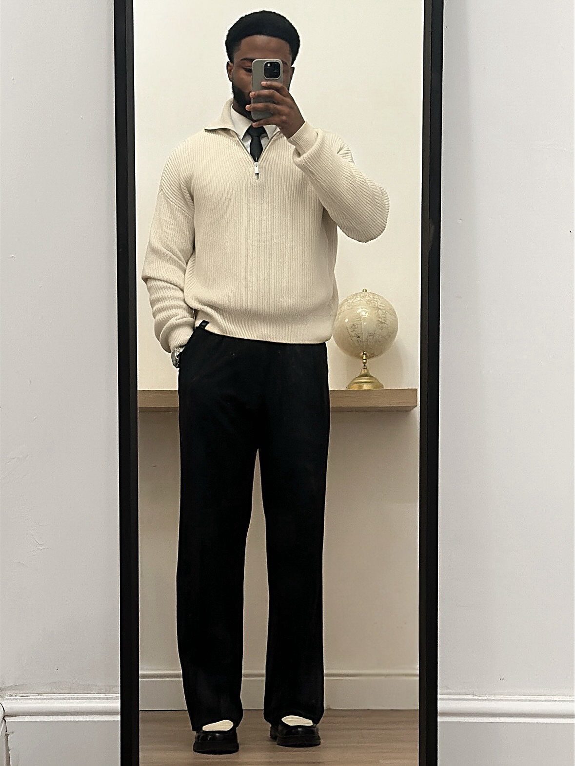 Man in a cream quarter-zip sweater, white shirt, tie, and black trousers posing for a mirror selfie.