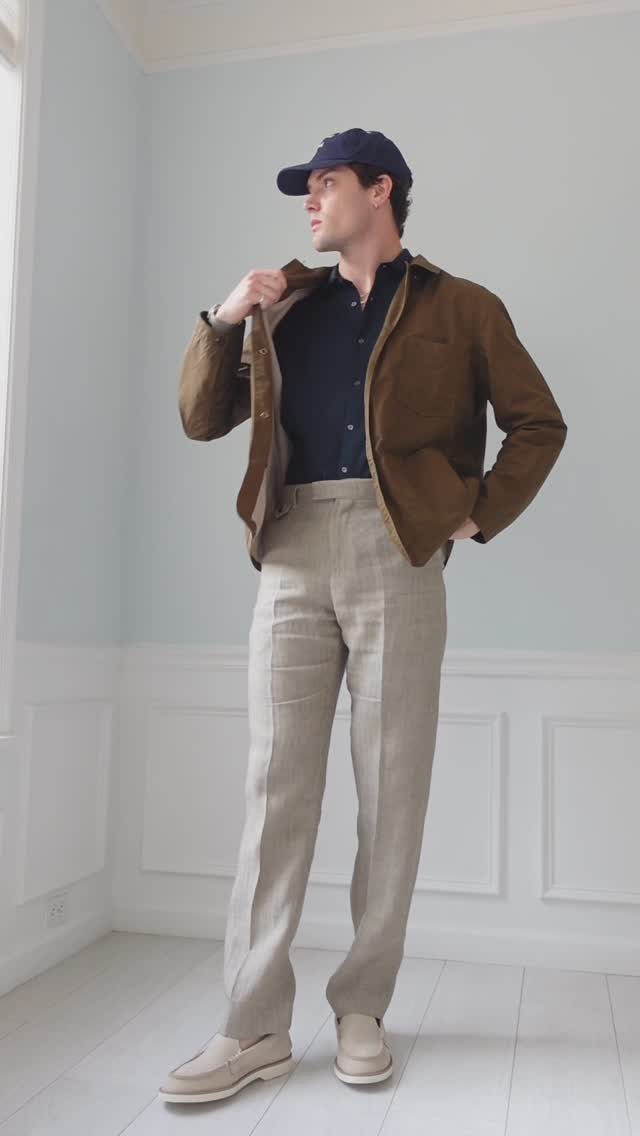 A man posing in a brown jacket, navy polo, beige trousers, and a navy cap.