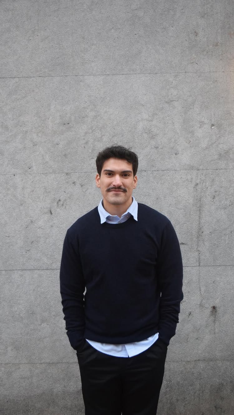 A man wearing a navy sweater and light blue collared shirt standing against a grey wall.