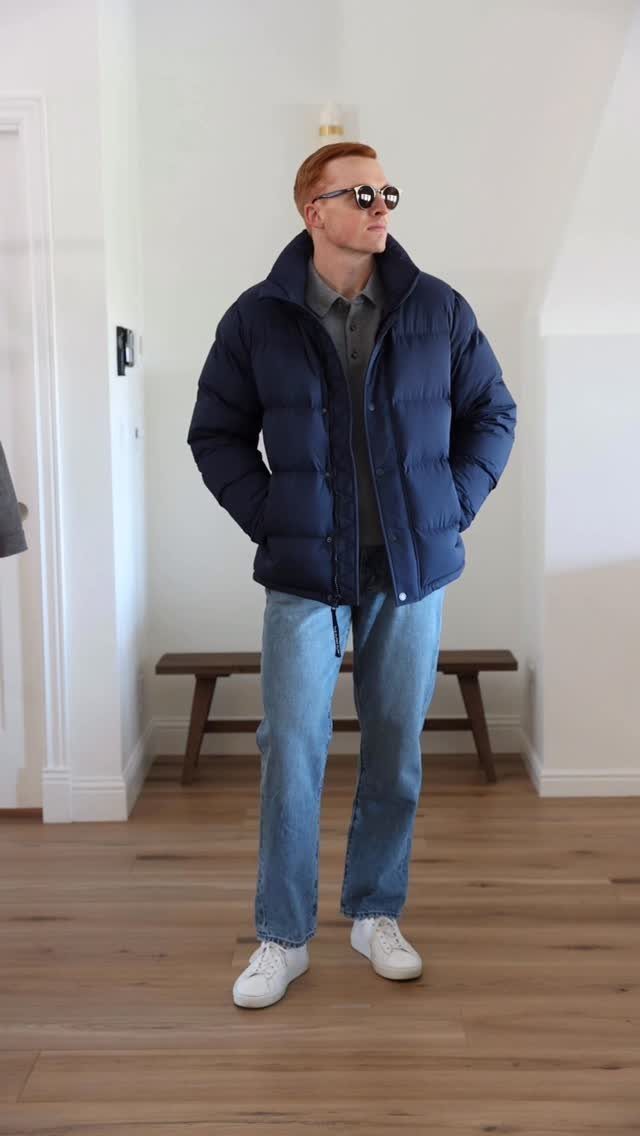 A man wearing a navy puffer jacket, grey polo, blue jeans, and white sneakers.