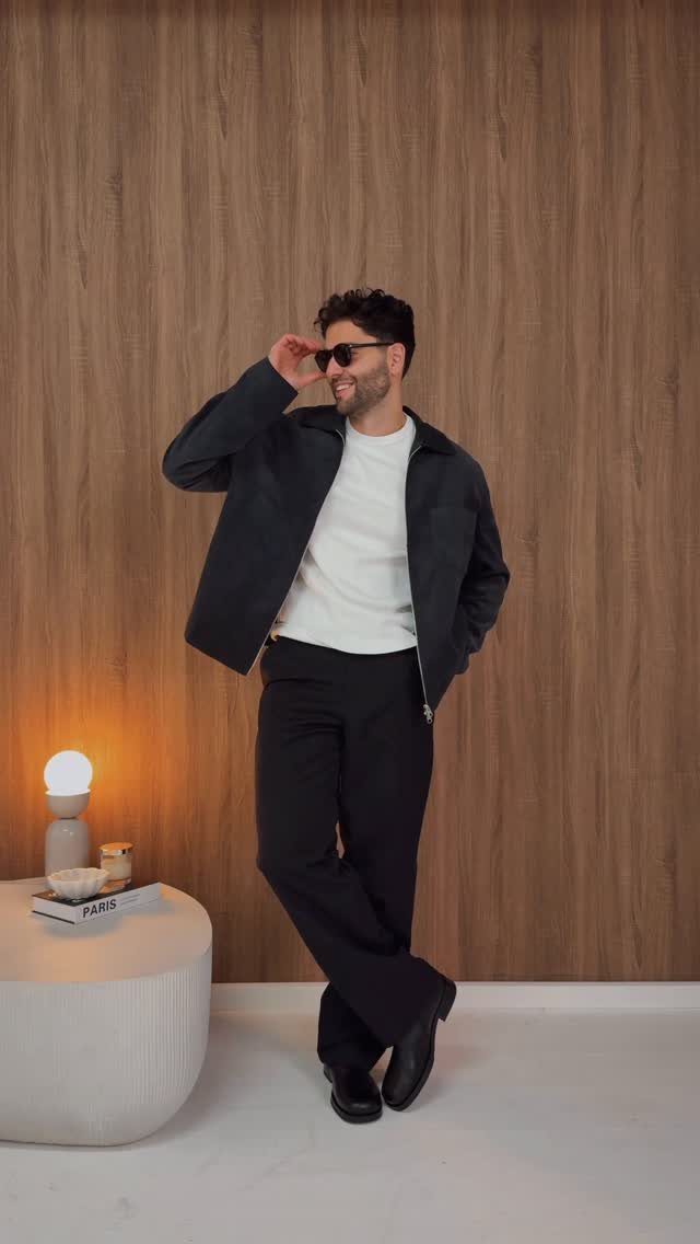 Man in a white t-shirt, dark jacket, and black trousers posing against a wood wall.
