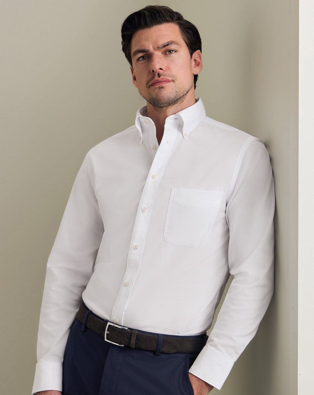 Man in a white button-down shirt and navy trousers leaning against a wall.