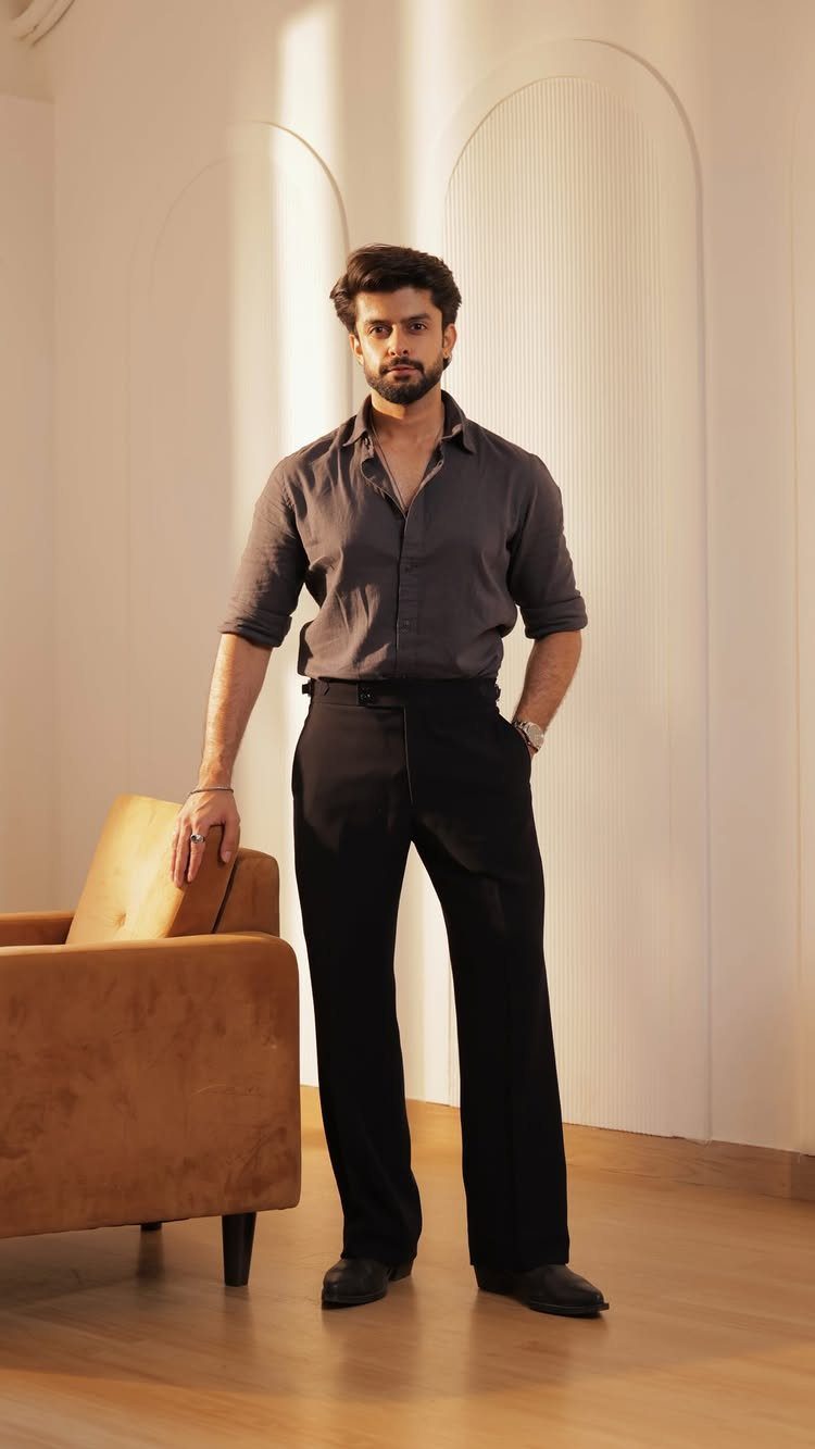 A man posing in a grey button-down shirt and black trousers next to an armchair.