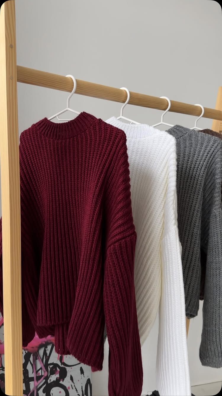 Three ribbed chunky knit sweaters in burgundy, white, and grey hanging on a wooden clothing rack.
