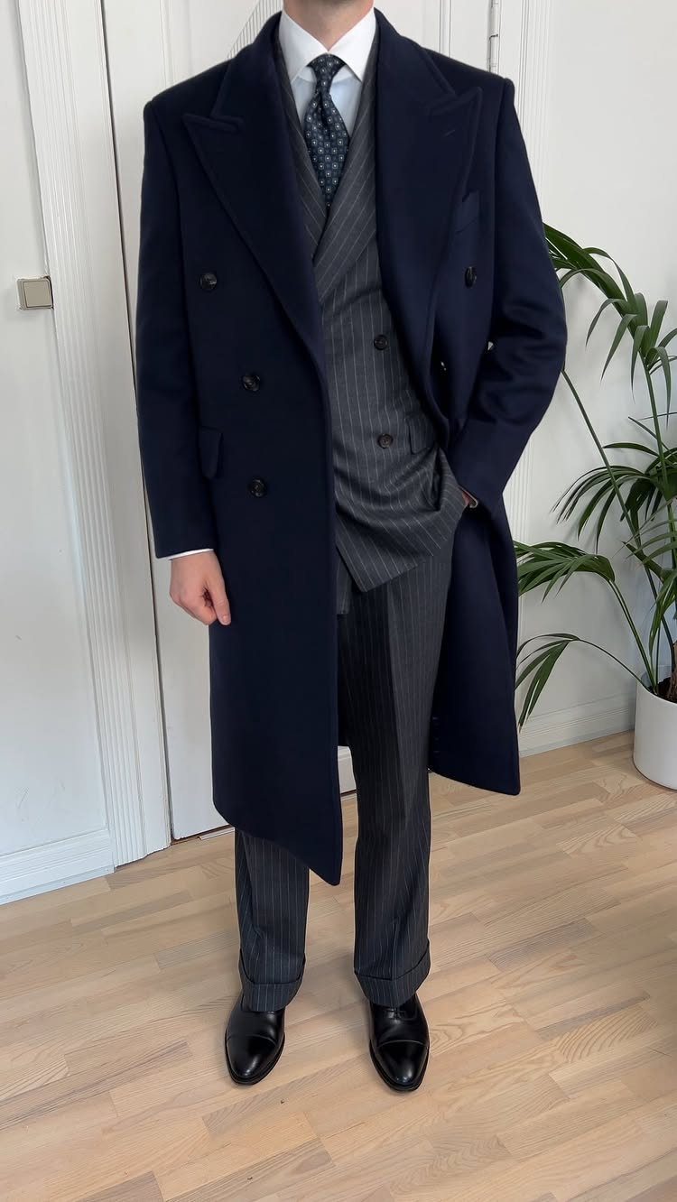 Man wearing a navy blue overcoat, grey pinstripe suit, and black dress shoes.