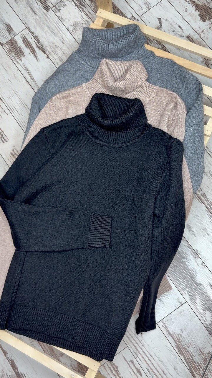 Three folded turtleneck sweaters in black, beige, and grey stacked on a wooden floor.