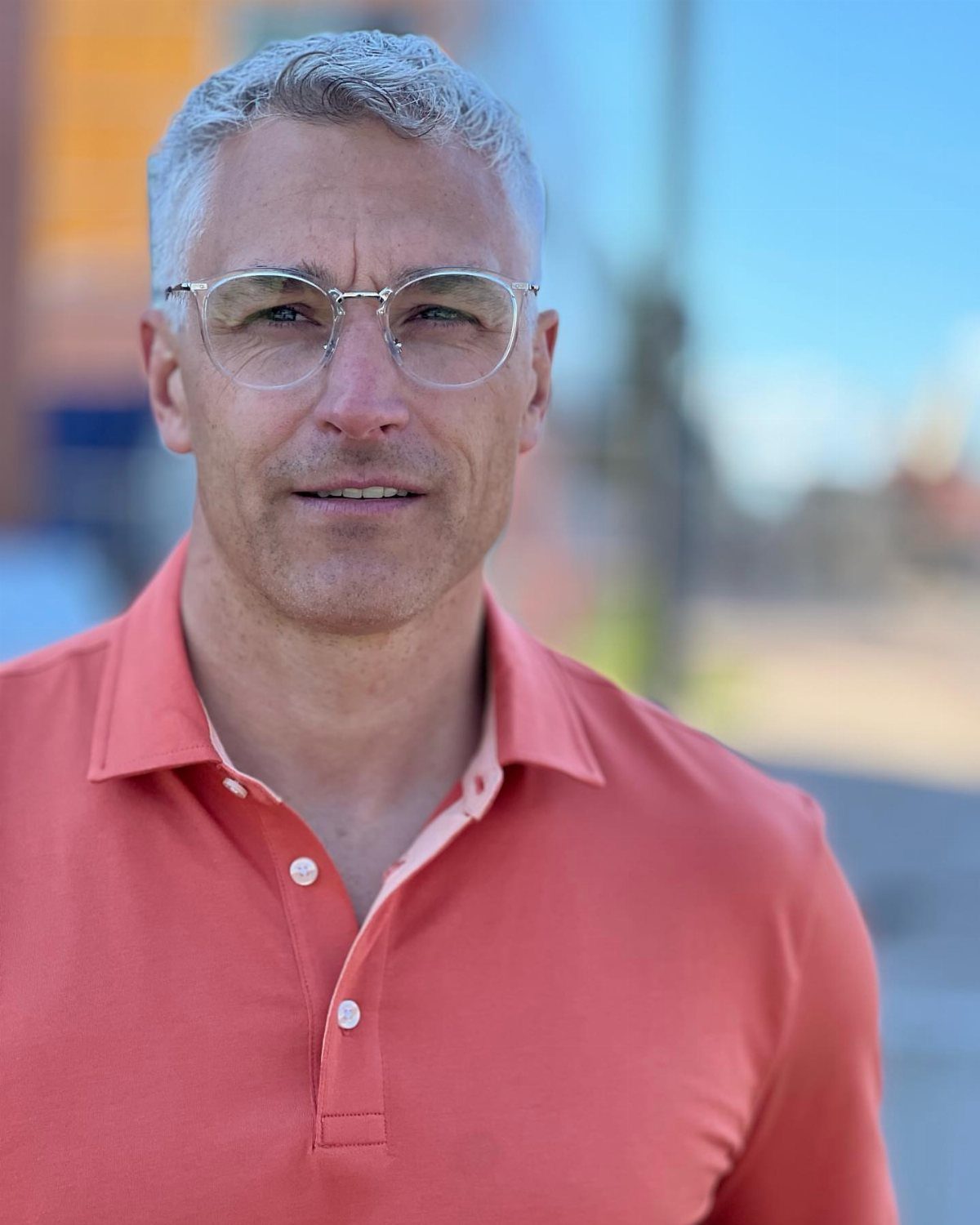 Middle-aged man with grey hair and glasses wearing a salmon polo shirt outdoors.