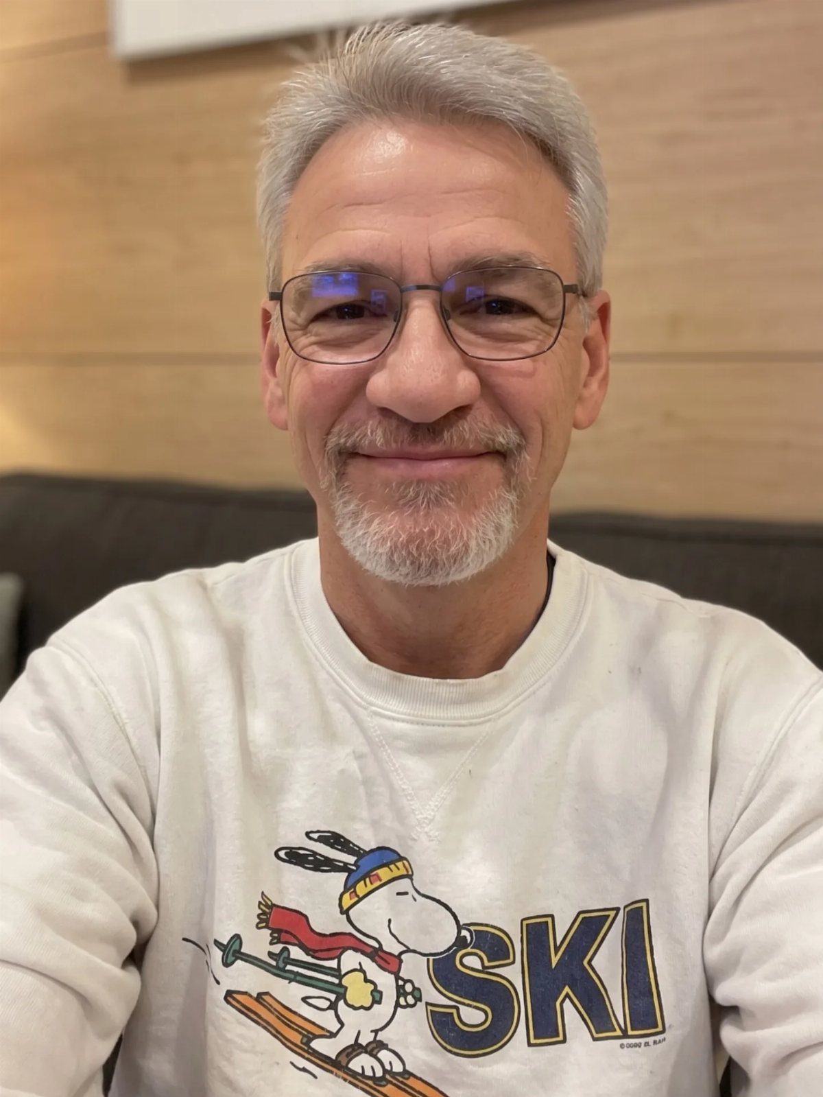 Smiling older man with grey hair and glasses wearing a white Snoopy graphic sweatshirt.