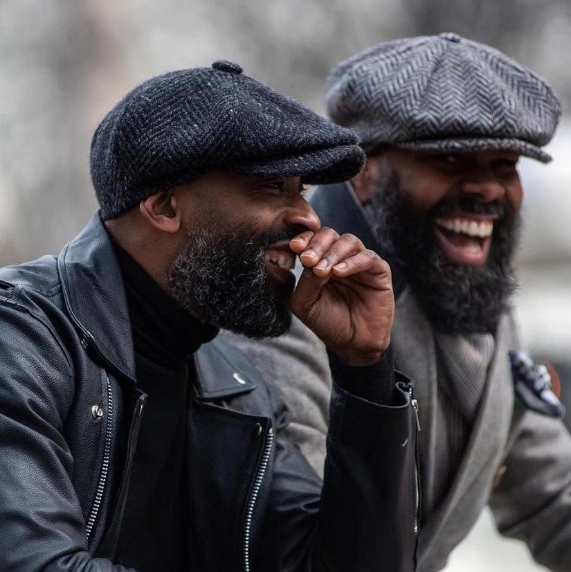 Two bearded men smiling while wearing stylish textured flat caps and jackets outdoors.