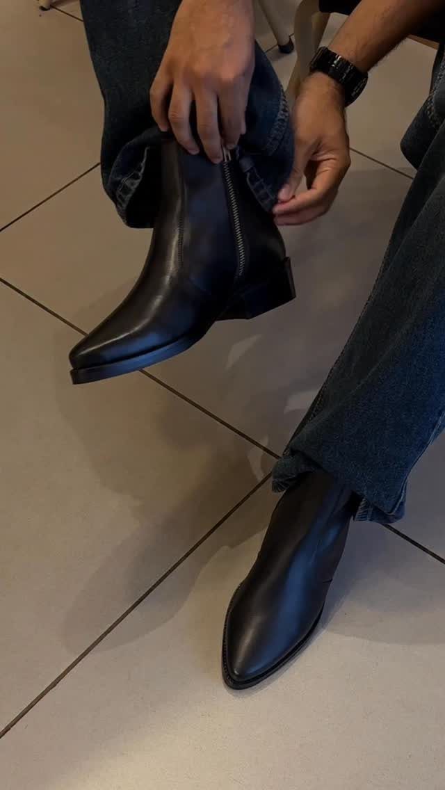 A person wearing dark denim jeans and black leather boots on a tiled floor.