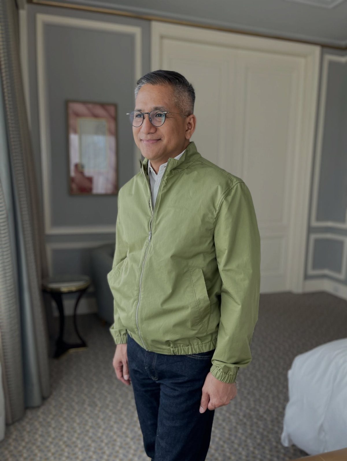A smiling middle-aged man wearing a green jacket and blue jeans standing in a room.