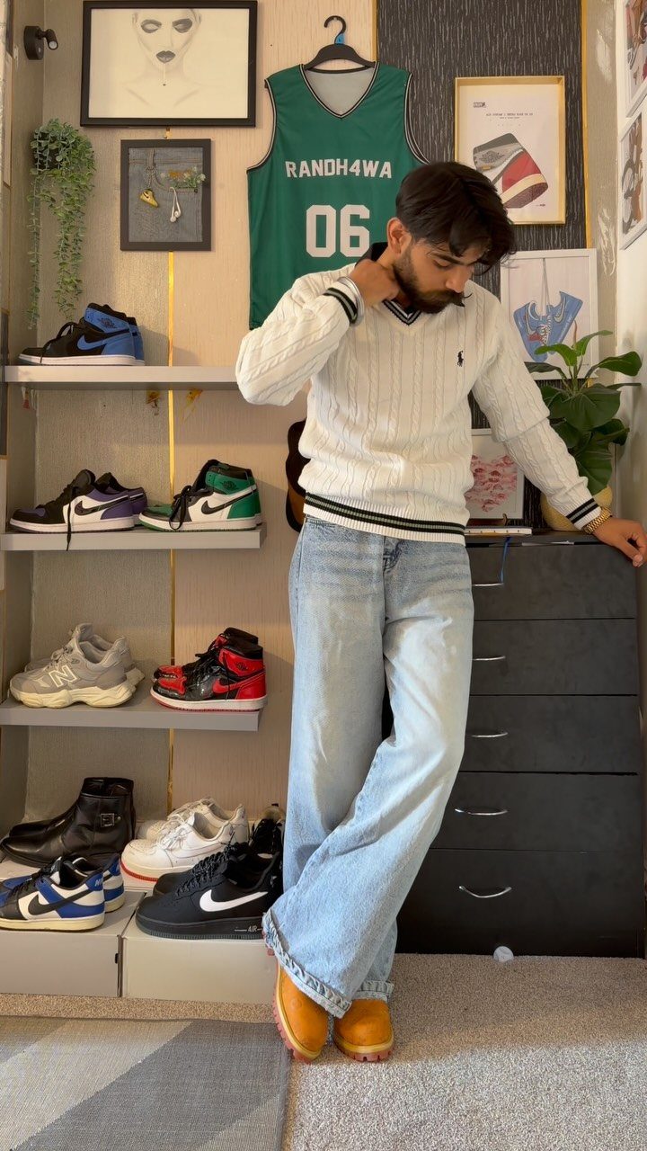 A man posing in a white cable-knit sweater, light blue jeans, and tan boots.