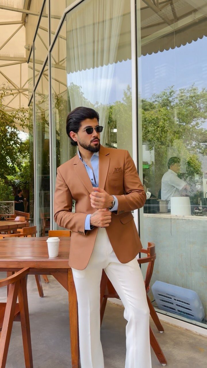 A man in a tan blazer, light blue shirt, and white trousers posing outdoors.