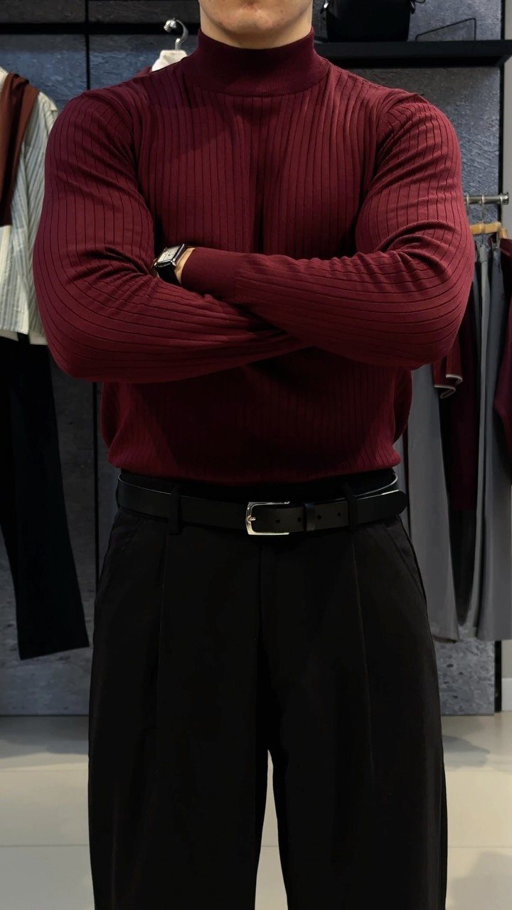 A person wearing a maroon ribbed turtleneck sweater and black trousers with a belt.