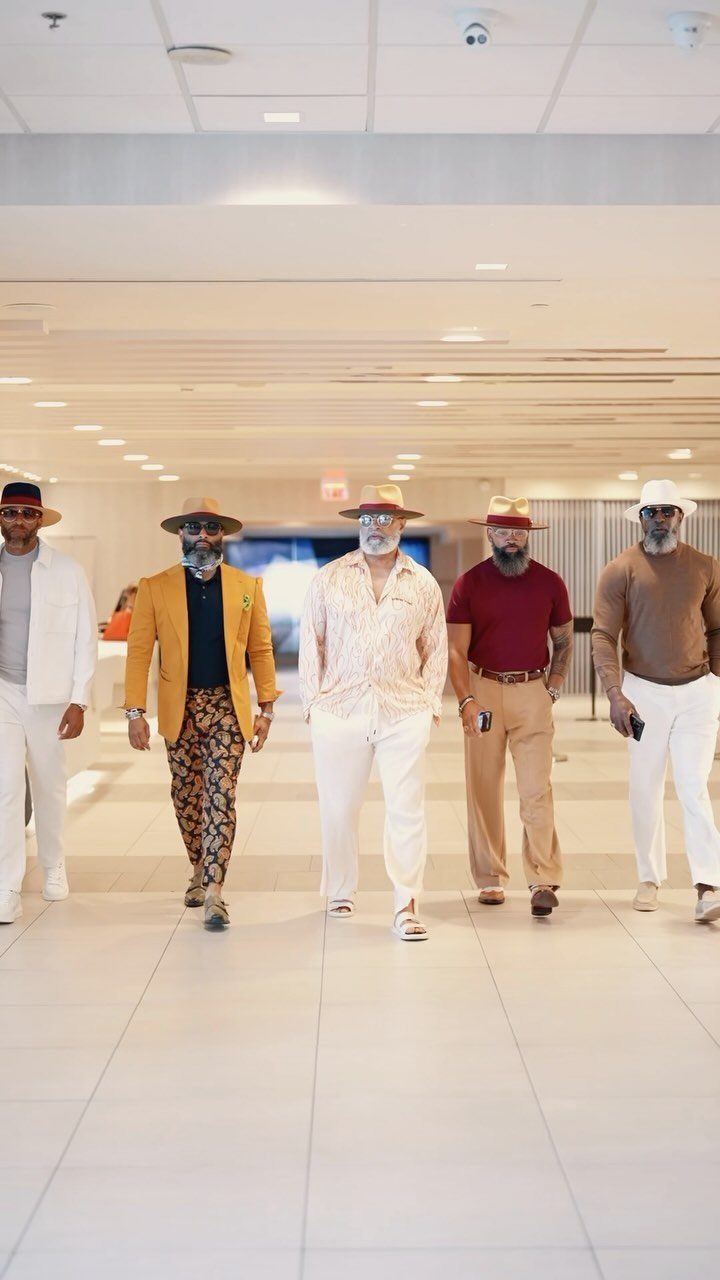 Five men walking in a hallway wearing stylish casual outfits and various hats.