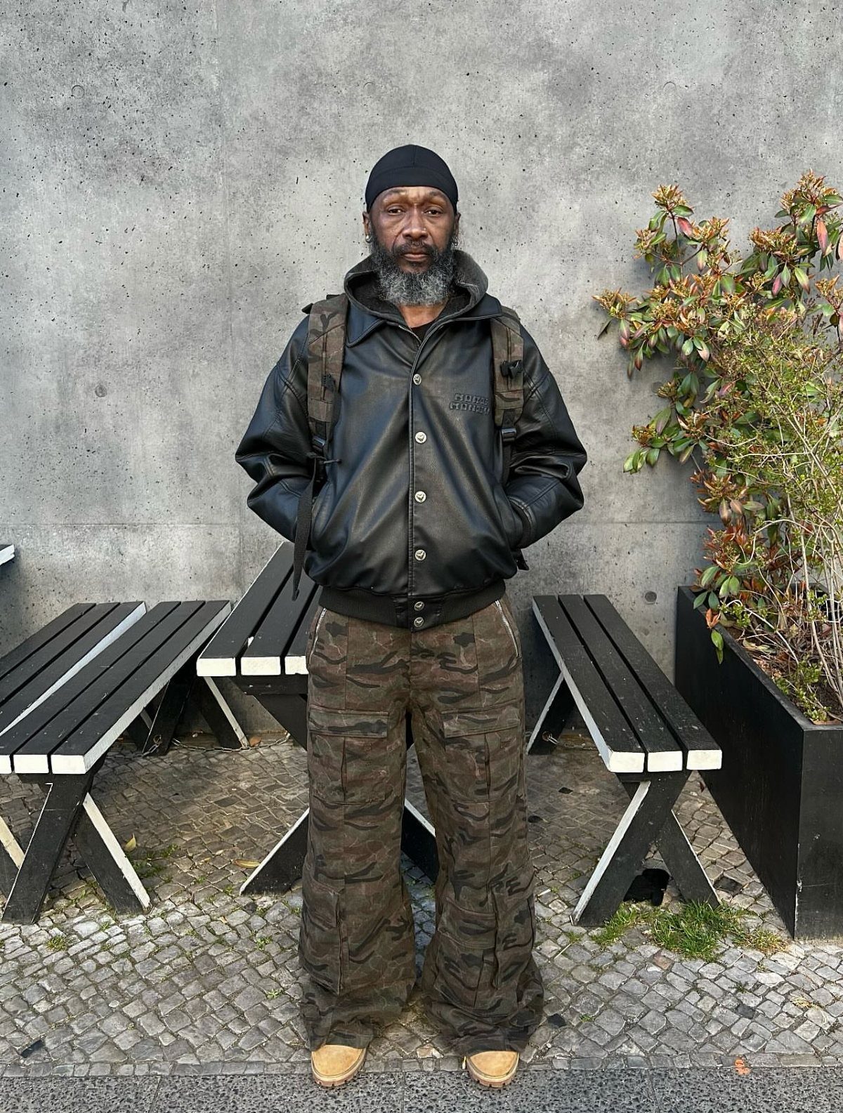 A bearded man wearing a black leather jacket, black beanie, and camouflage cargo pants standing outdoors.